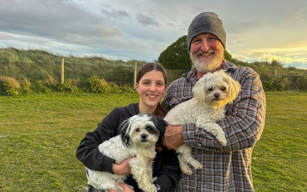 An older man in a beanie and flannel shirt with a trimmed beard holds a small white dog. Next to him a young girl with a brown ponytail holds a small black and white dog. The pair are standing in a grassy field underneath a cloudy sky.