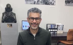 David sits at a desk with a large, old, ornate book in front of him. He smiles at the camera.