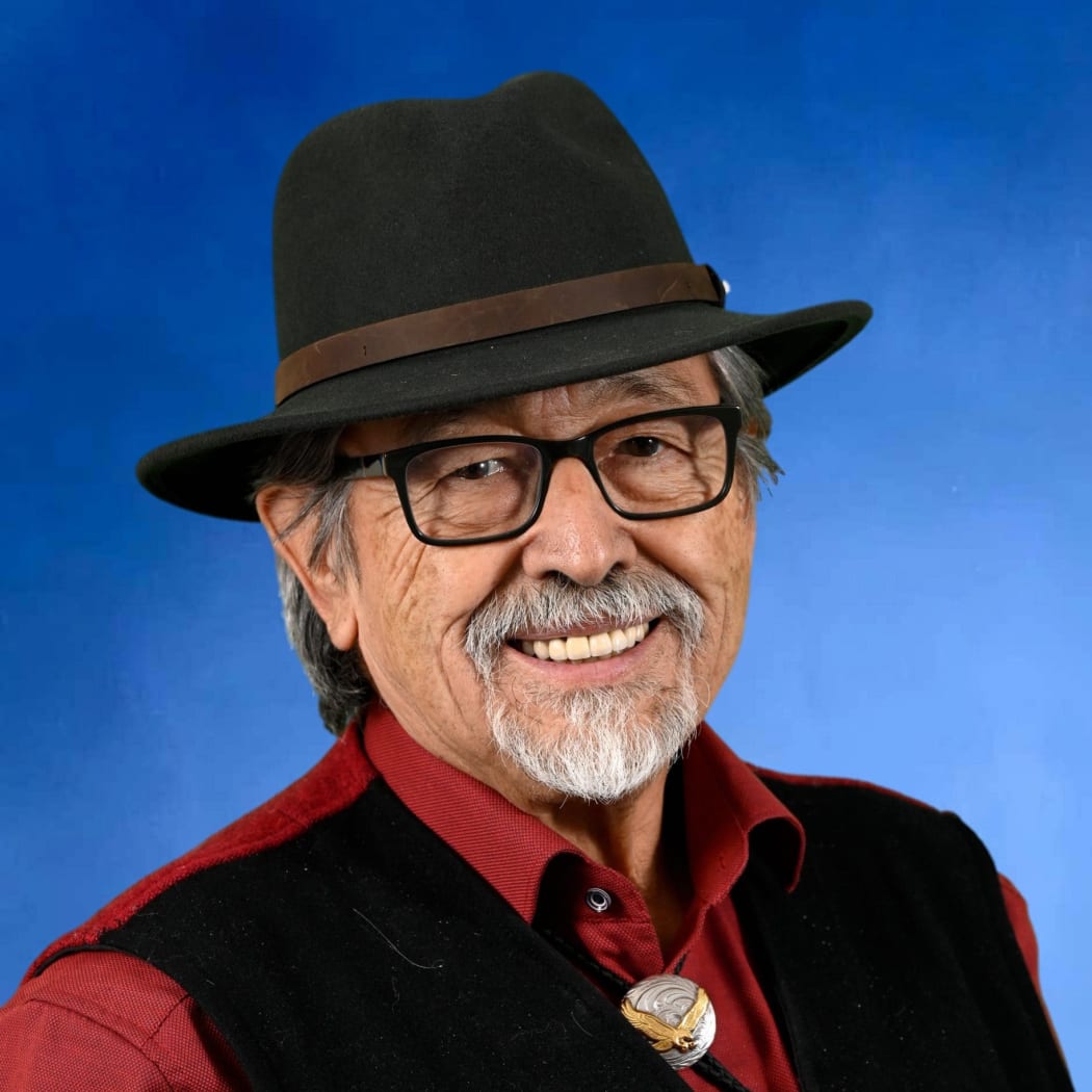 Ron smiles at the camera. He wears a hat, glasses, and shirt with a bolo tie.