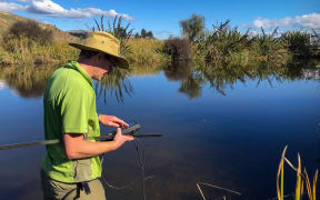 A man wearing a wide-brimmed hat and a bright green colllared t-shirt stands on the edge of a wetland pool framed by long grass and vegetation. He is side on, holding a long pole and using an electronic device with a wire extending down towards the water. It's a sunny day and the water is still.