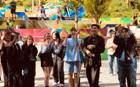 A group of people are gathered in a civic square, dancing, clapping and cheering.