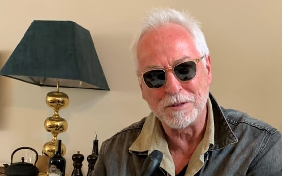 Eddie Raynor holding a microphone at a kitchen table, a lamp and teapot in the background.