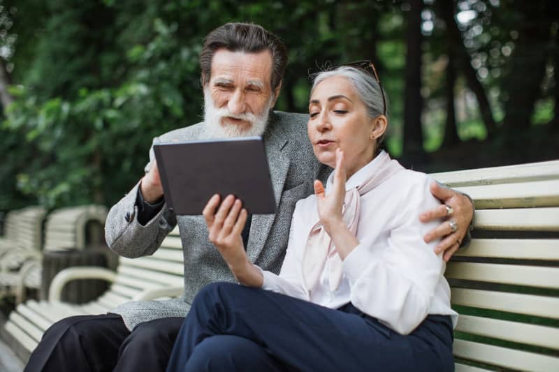 An elderly couple sit on a park bench speaking into an iPad.