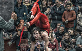 Shannen is doing a handstand and firing an arrow with her feet. There is a crowd behind her.