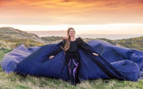 Toni Huata stands on top of a mountain wearing a purple cape that is billowing in the wind.