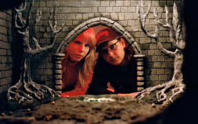 Two people - one with long blonde hair and bangs, one wearing a cap and glasses - peer into the lens through the entrance of a sculpture of a medieval castle.
