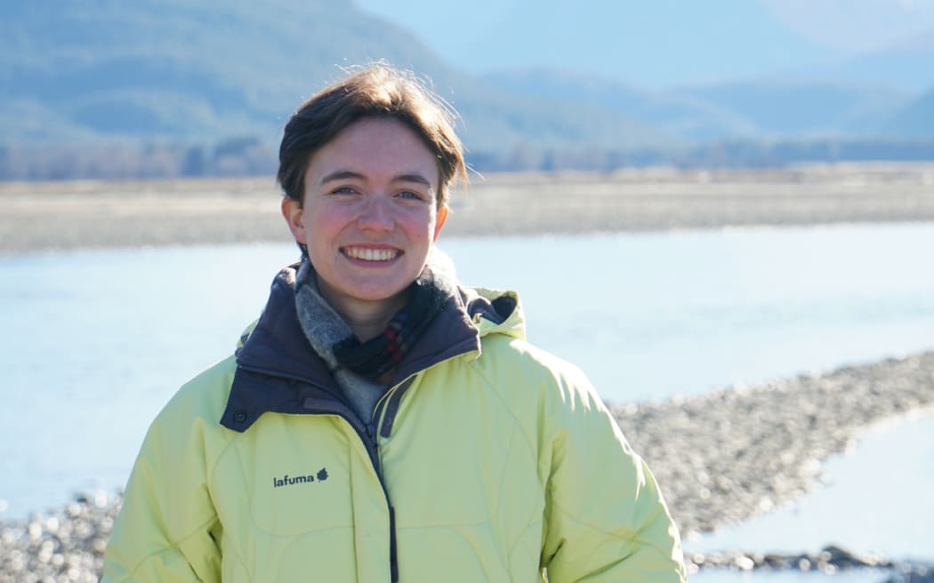 Carmen stands in front of a river wearing a puffer jacket. She smiles. Mountains rise into the distance.