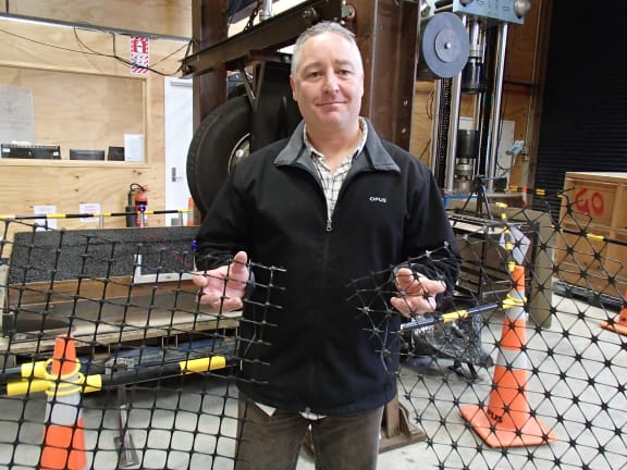 Steve Bagshaw is holding different kinds of geogrids, which are being tested with the accelerated pavement wear machine behind to see how effective they are at creating a consolidated base course of rounded river gravels.The base course is the load bearing part of the road.