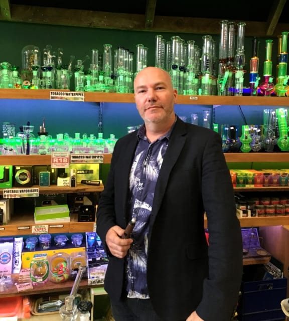A portrait of Hemp Store owner and NORML NZ President Chris Fowlie in fromt of a range of pipes and vaporisers.
