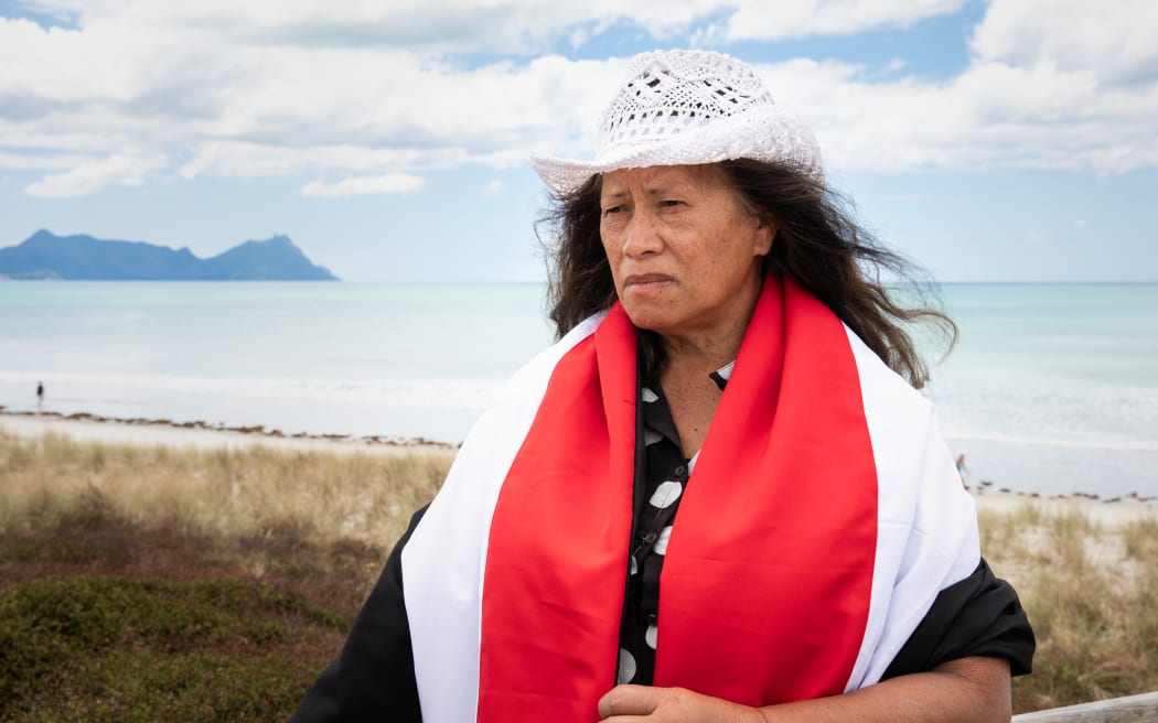 Josephine Pirihi stands on the sands of Ruakaka Beach with a red, white, and black banner - the colours of the Tino Rangatiratanga flag - cloaked around her shoulders.