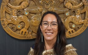 Claire Charters is wearing glasses with long brown hair, she is standing up and smiling at the camera.