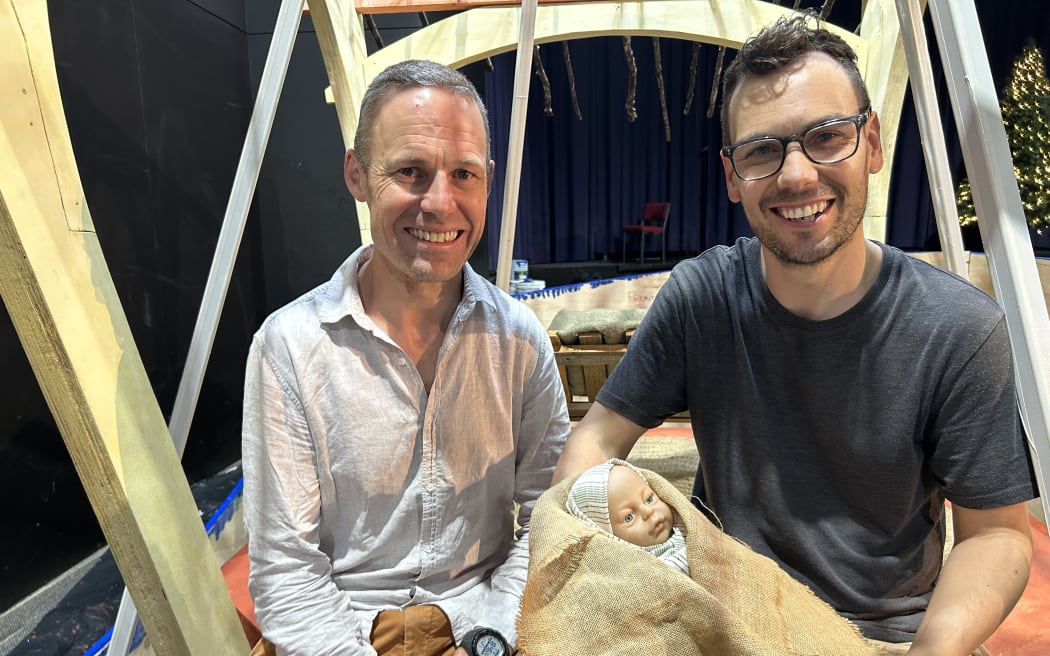 Photo of two men sitting down, with one person cuddling a doll as baby Jesus.