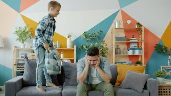 A young boy jumps on a couch with a cushion next to a man with his hands covering his ears.