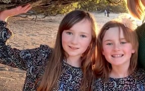 Two sisters, probably Pākehā, aged 8 and 10 in matching navy-blue frocks with a busy floral pattern, smiling at camera. One has long brown hair the other possibly shoulder-length or longer but hidden behind her back.