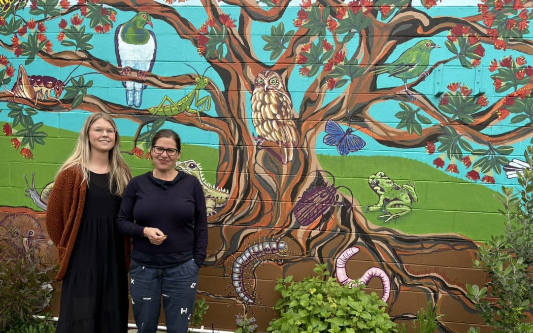 Two smiling women standing in front of a mural of a tree with creatures in its branches and on its trunk and the soil around it.