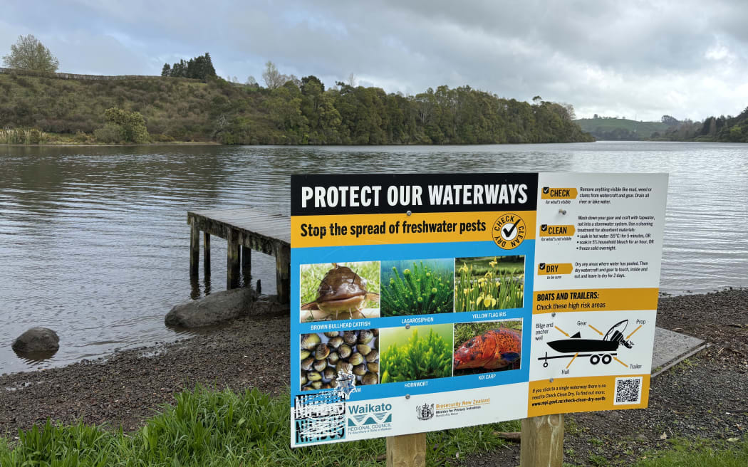 In the foreground is a large sign saying 'Protect Our Waterways - stop the spread of freshwater pests'. Under that are images of the pests of Karapiro lake - catfish, koi carp, gold clam, and three different weeds. The check clean dry checklist is beside it.