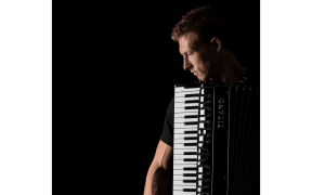 Side view of accordionist Grayson Masefield. He holds the accordion vertically, with the keyboard immediately below his head.