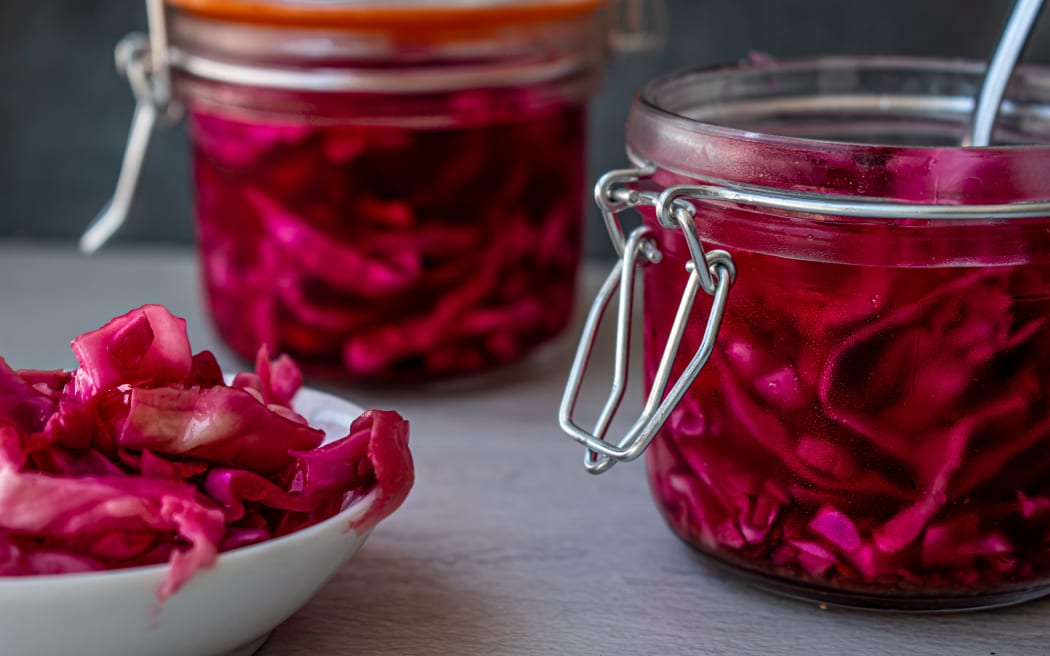 Image contains two jars of red cabbage sauerkraut, with a small pile of kraut in a white dish in the foreground.
