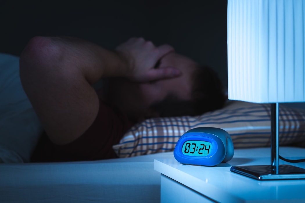 Insomnia and sleepless concept. Man unable to sleep. Exhausted and tired. Covering face with hand. Alarm clock on nightstand and bed in bedroom.