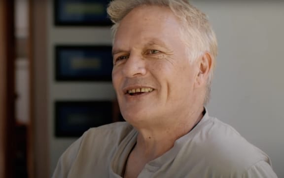 A smiling white-haired man in a grey t-shirt looks to the right of camera.