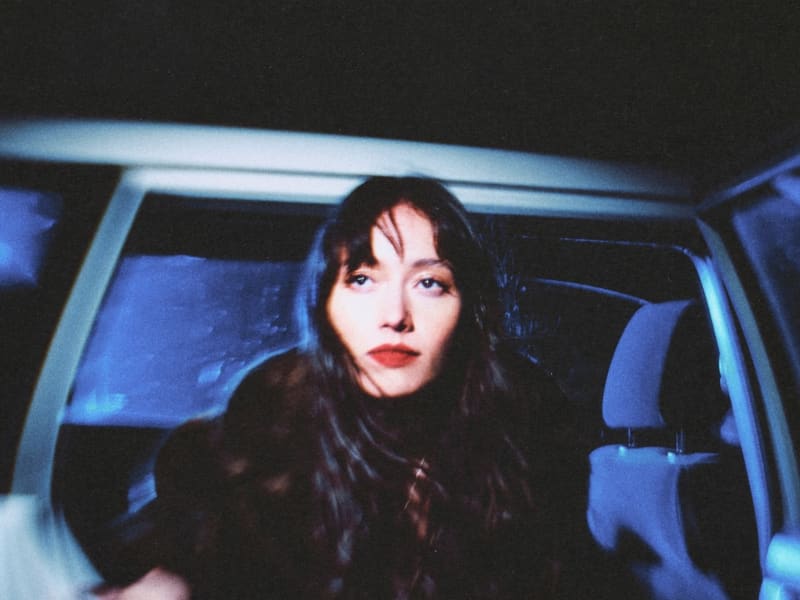 Fazerdaze - a young woman with long dark hair and lipstick - looks out from an open car door.
