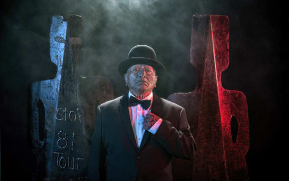 Image from Tame Iti: I Will Not Speak Māori Exhibition.  Artist wearing a suit, bowtie and bowler hat stands in front of metal sculptures in a stylised human form.
