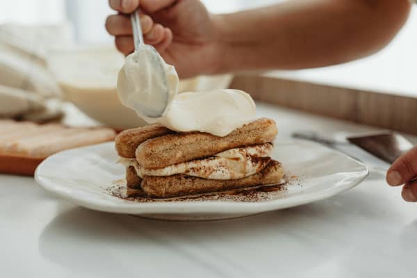 A person dolloping on freshly whipped cream on finger biscuits to make a tiramisu.
