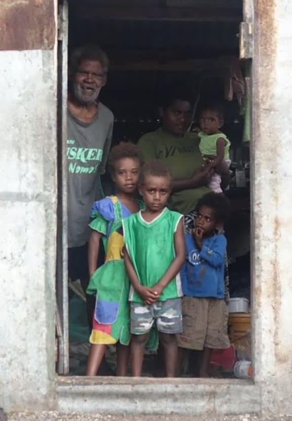 Grandfather Sakaio Cook stands in the doorway of his house, one of only two on Mataso that survived cyclone Pam, with daughter Leimas carrying baby Margaret, Rexson in the green shirt, Ellen in the blue shirt and Leika in the vanuatu dress(green yellow and red).