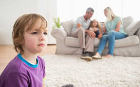 Photo of an upset boy sitting on floor while parents enjoying with sister on sofa at home