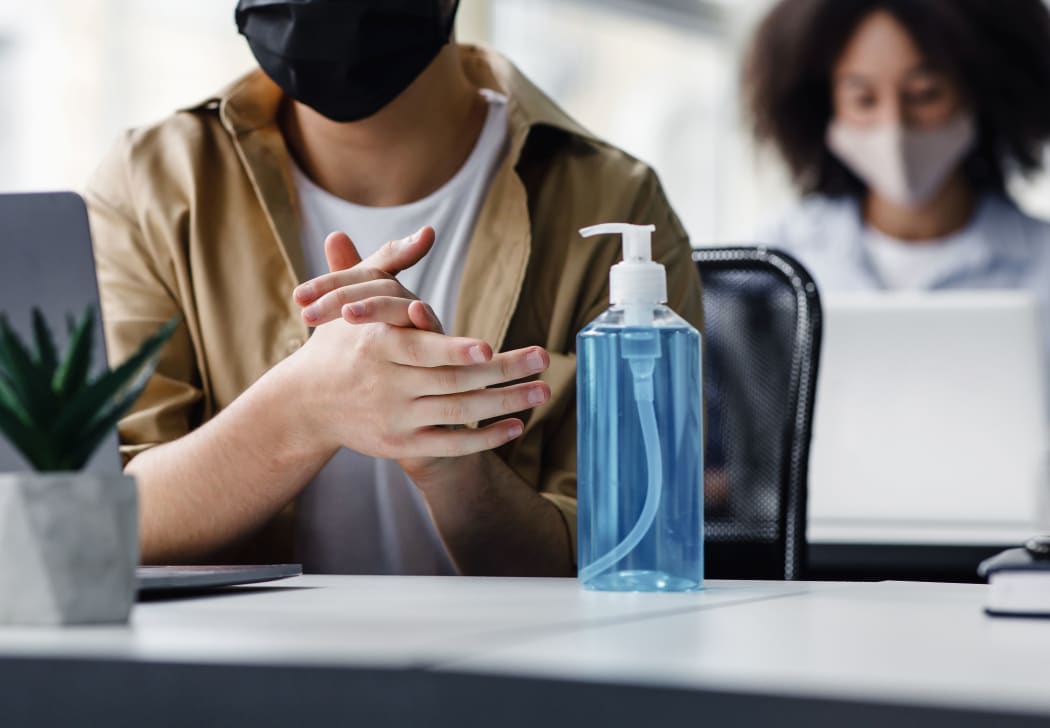 Focus on antiseptic to protect against covid virus. Guy disinfects his hands, sitting at table at workplace with laptop, working in interior of modern office, panorama, copy space