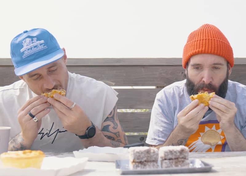 Two men wearing hats sit outside eating pies.