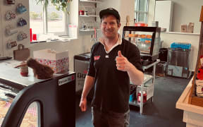 A man dressed in a black rugby jersey and cap stands in the kitchen of a pie shop. The tino rangatiratanga flag is hung up in the background.