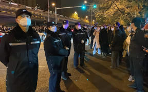 Policemen watch over protesters gathering along a street during a rally for the victims of a deadly fire as well as a protest against China's harsh Covid-19 restrictions in Beijing on November 28, 2022. - A deadly fire on November 24, 2022 in Urumqi, the capital of northwest China's Xinjiang region, has become a fresh catalyst for public anger, with many blaming Covid lockdowns for hampering rescue efforts, as hundreds of people took to the streets in China's major cities on November 27, 2022 to protest against the country's zero-Covid policy in a rare outpouring of public anger against the state. Authorities deny the claims. (Photo by Michael Zhang / AFP)