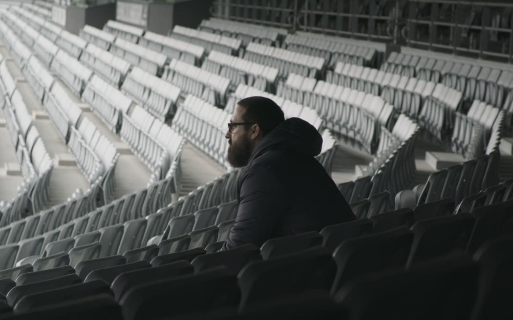 Luke sits in an empty stadium seating section, looking out at the field.