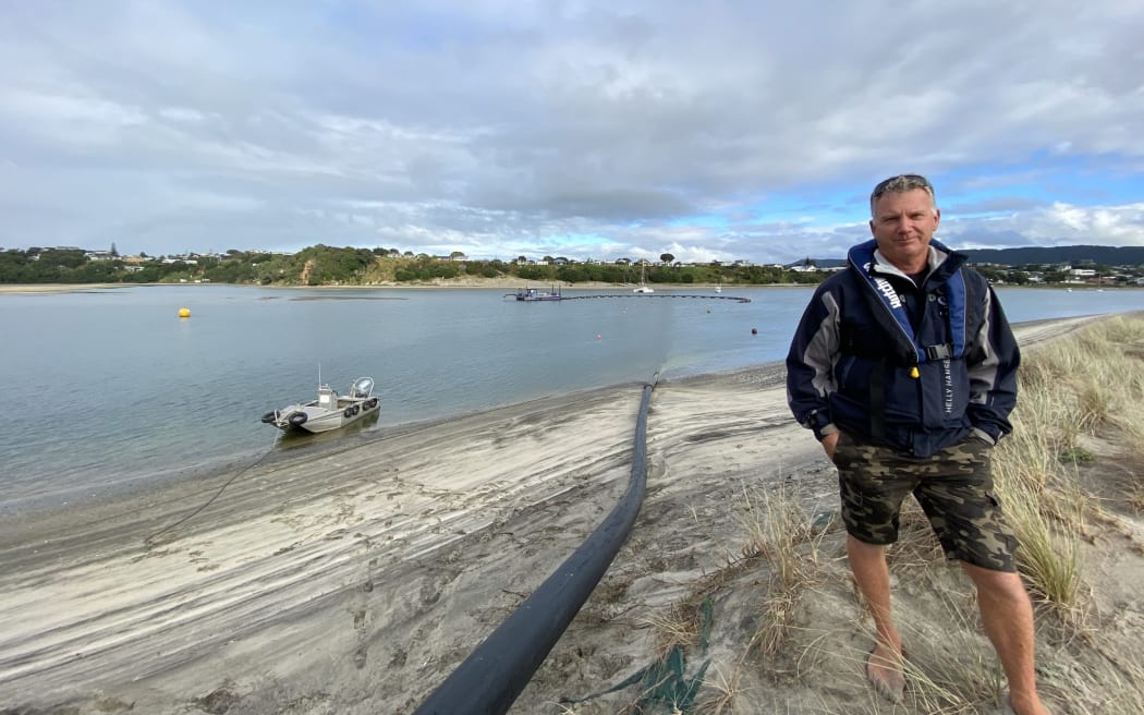 Mangawhai Harbour Restoration Society dredge operator Mark Vercoe on the sandspit where the barge in the background pumps sand along the black pipe beside him, to keep the harbour open and build up the protective sand dune barrier known as a bund wall that protects the sandspit from harbourside-generated erosion.