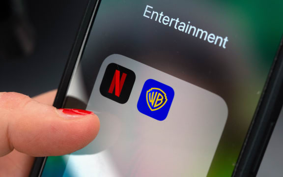 A finger taps an app folder field on a smartphone with the logos of the streaming platform Netflix (L) and the film studio Warner Brothers.