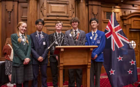 Five students in school uniforms pose for a picture behind a wooden rostrum in Parliament's debating chamber.