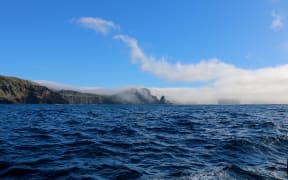 An island with rugged steep cliffs and tussock vegetation extends from the left, with another smaller outcrop/island off to the right. Both are cloaked with low cloud and mist, but the sky is blue above. The ocean in the foreground is deep blue and choppy.
