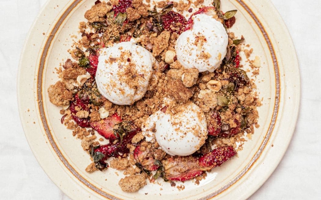 A birds' eye view of a beige plate on a white table. The plate is strewn with crumble, chunks of strawberry, and three lumps of ice cream.