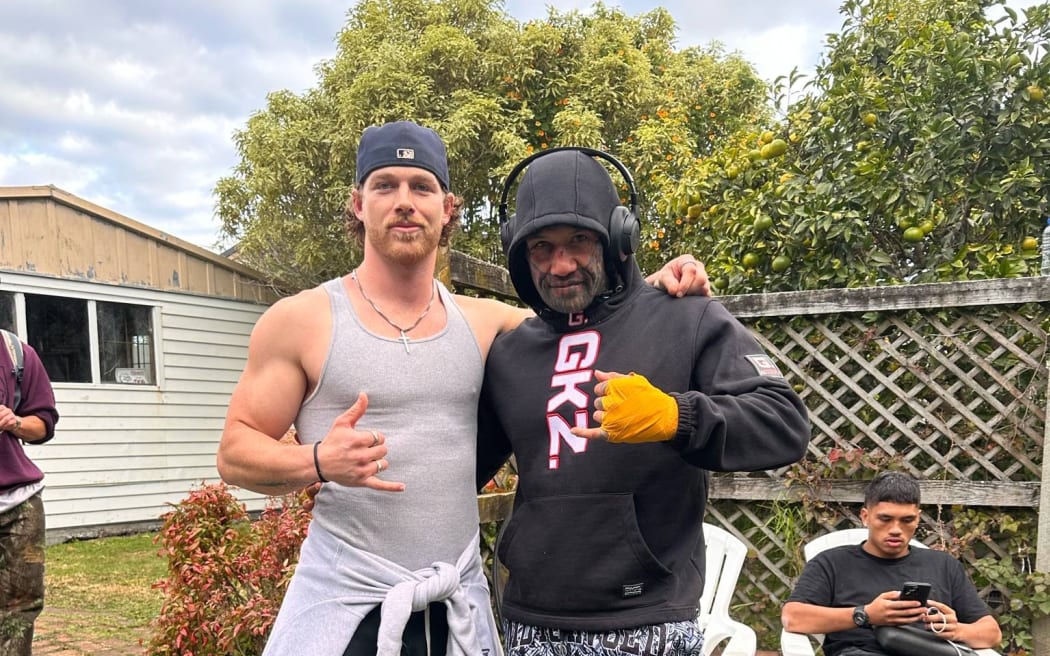 Two men standing on a lawn in what appears to be a back garden. There is a fence and a white garage in the background.