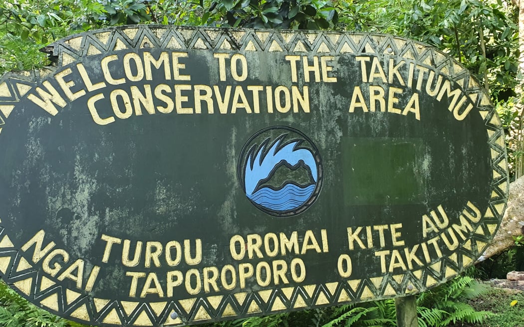 A large green oval sign in front of a forest background. In yellow are the letters 'Welcome to the Takitumu Conservation Area' on the top and 'Tauou Oromai Kite Au Ngai Taporoporo O Takitumu' on bottom, with a logo in between.