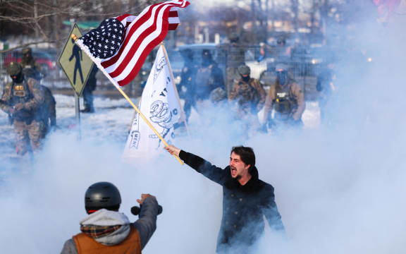 A man waves a US flag next to people protesting US Immigration and Customs Enforcement (ICE) near the Bishop Henry Whipple Federal Building in Minneapolis, Minnesota, on January 9, 2026. A US Immigration and Customs Enforcement (ICE) agent shot and killed an American woman on the streets of Minneapolis January 7, leading to huge protests and outrage from local leaders who rejected White House claims she was a domestic terrorist. The woman, identified in local media as 37-year-old Renee Nicole Good, was hit at point blank range as she apparently tried to drive away from agents who were crowding around her car, which they said was blocking their way. (Photo by CHARLY TRIBALLEAU / AFP)