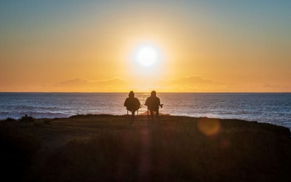 A couple sit together on fold out chairs watching a sunset over the sea.