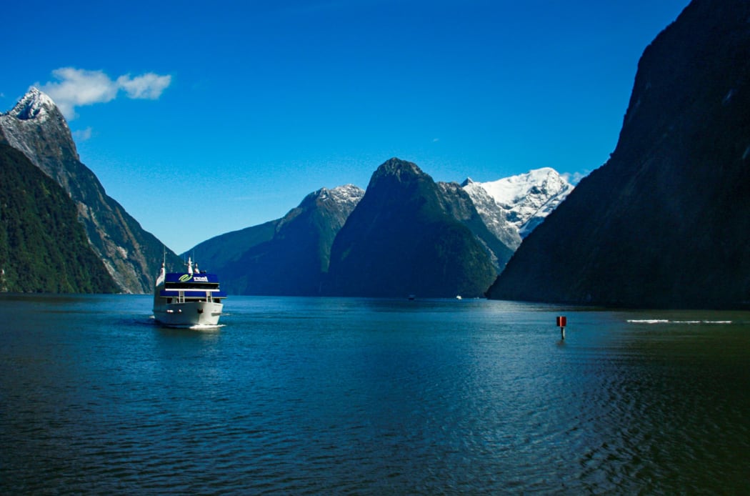 A tourist sightseeing ferry boat travels down the calm, deep blue waters of Milford Sound in the remote Fiordland region in the south west of the South Island of New Zealand