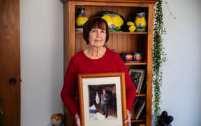 Image of woman (Julie Appelgren) standing in her home wearing a red long sleeved shirt, holding a framed photograph of her husband Ross Appelgren.