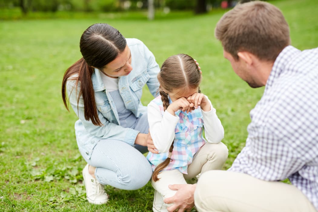 A photo of a little girl crying while sitting on grass and her parents comforting her during the weekend