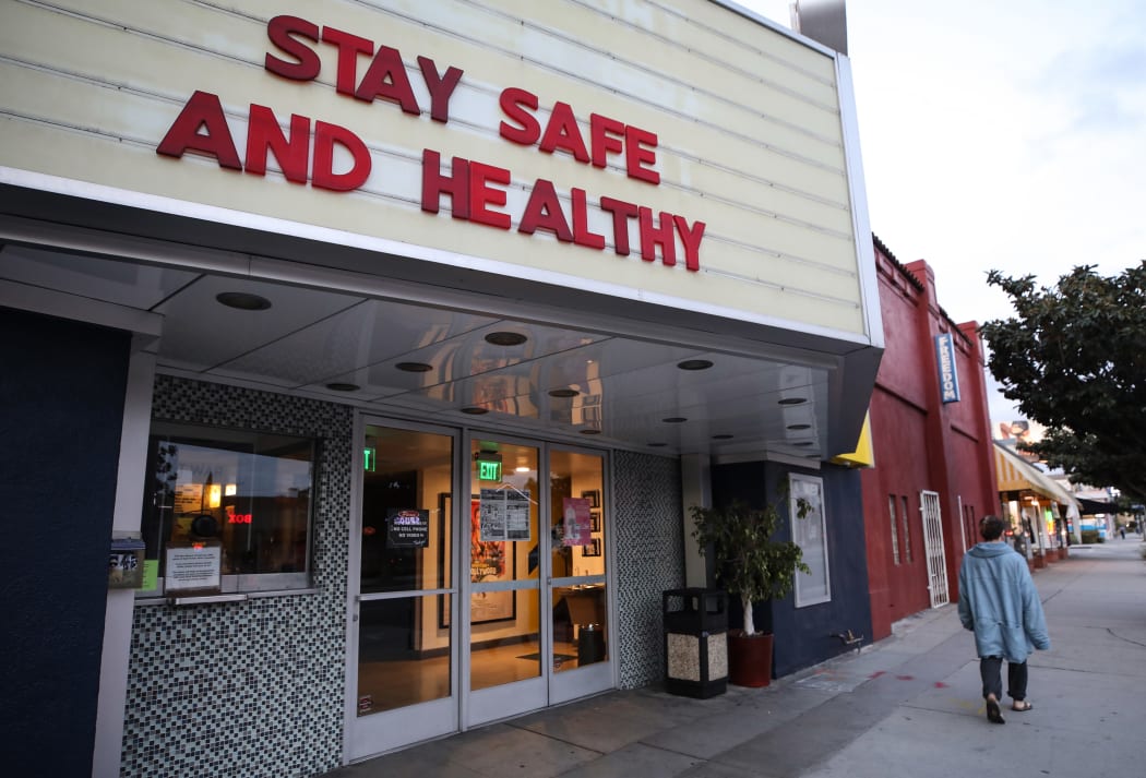 A pedestrian walks past a shuttered movie theater, with the message 'Stay Safe and Healthy' displayed on the marquee.