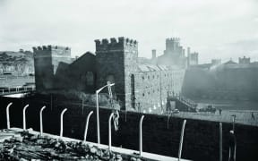 Mount Eden Prison on fire during rioting in 1965. In the foreground, Auckland Electric Power Board staff erect floodlights.