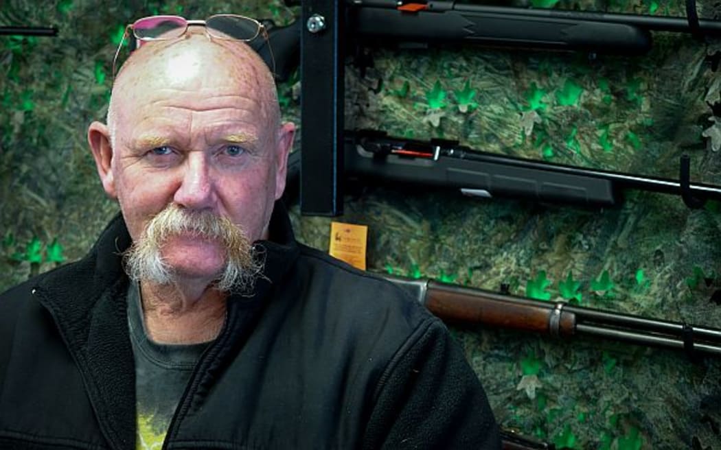 Man with impressive moustache stands in from of shop gun rack