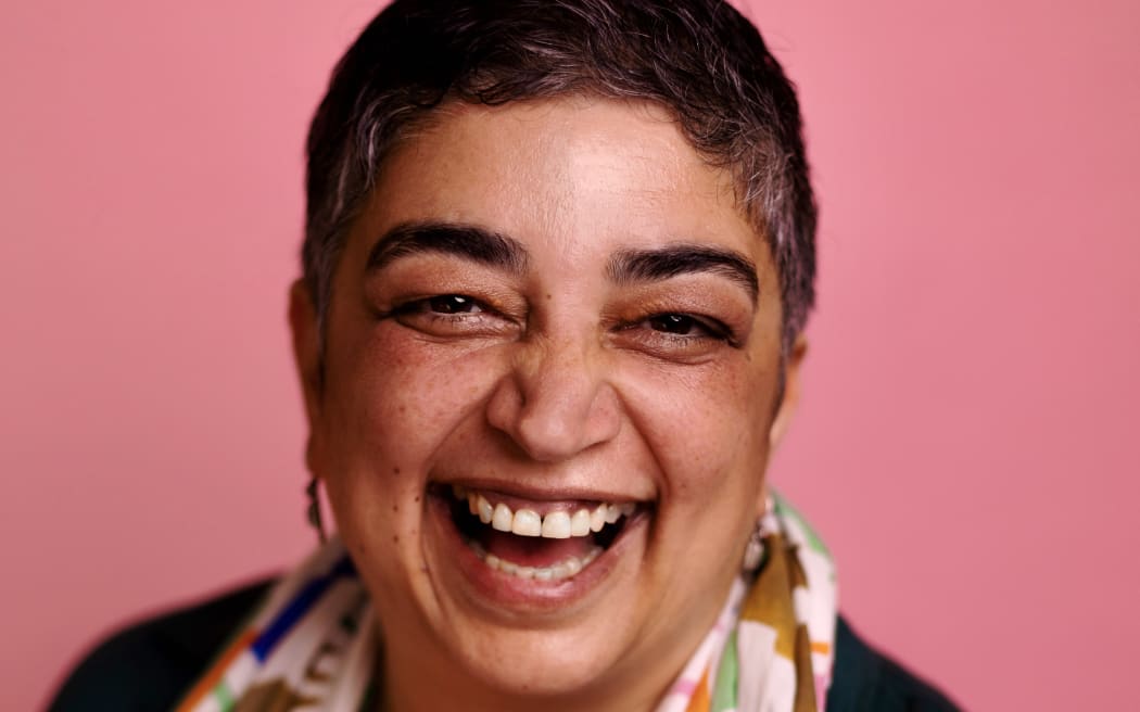 A photo of award winning storyteller, comedian, writer, director and political satirist Sameena Zehra. Sameena is laughing and wearing a multi-coloured scarf on a pink background.